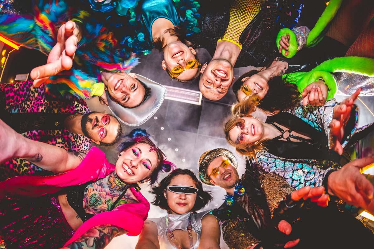 Eine Gruppe fröhlicher Menschen mit bunten Festival-Outfits und -Accessoires von Deiters