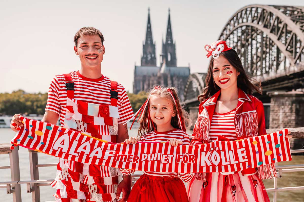 3 Personen vor der Kölner Skyline mit dem Kölner Mottoschal 2025/2026 und in rot-weißen Kostümen