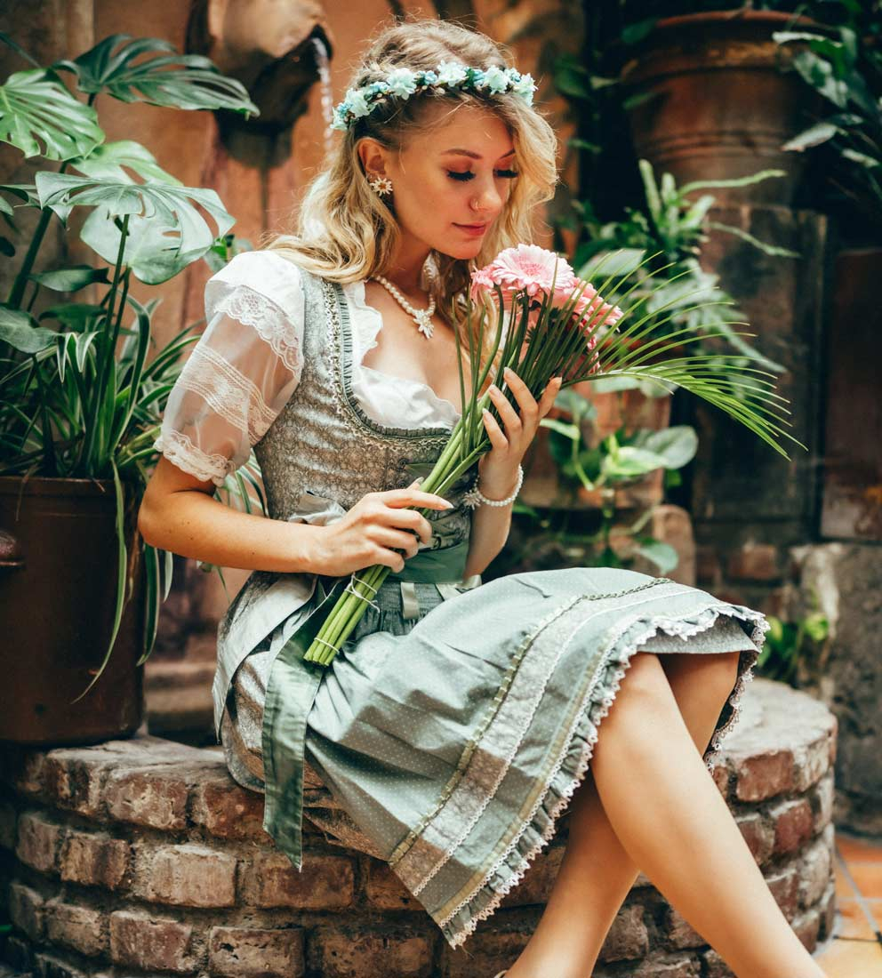 Eine Dame in mintfarbenem Dirndl sitzt am Brunnen mit Blumen in der Hand