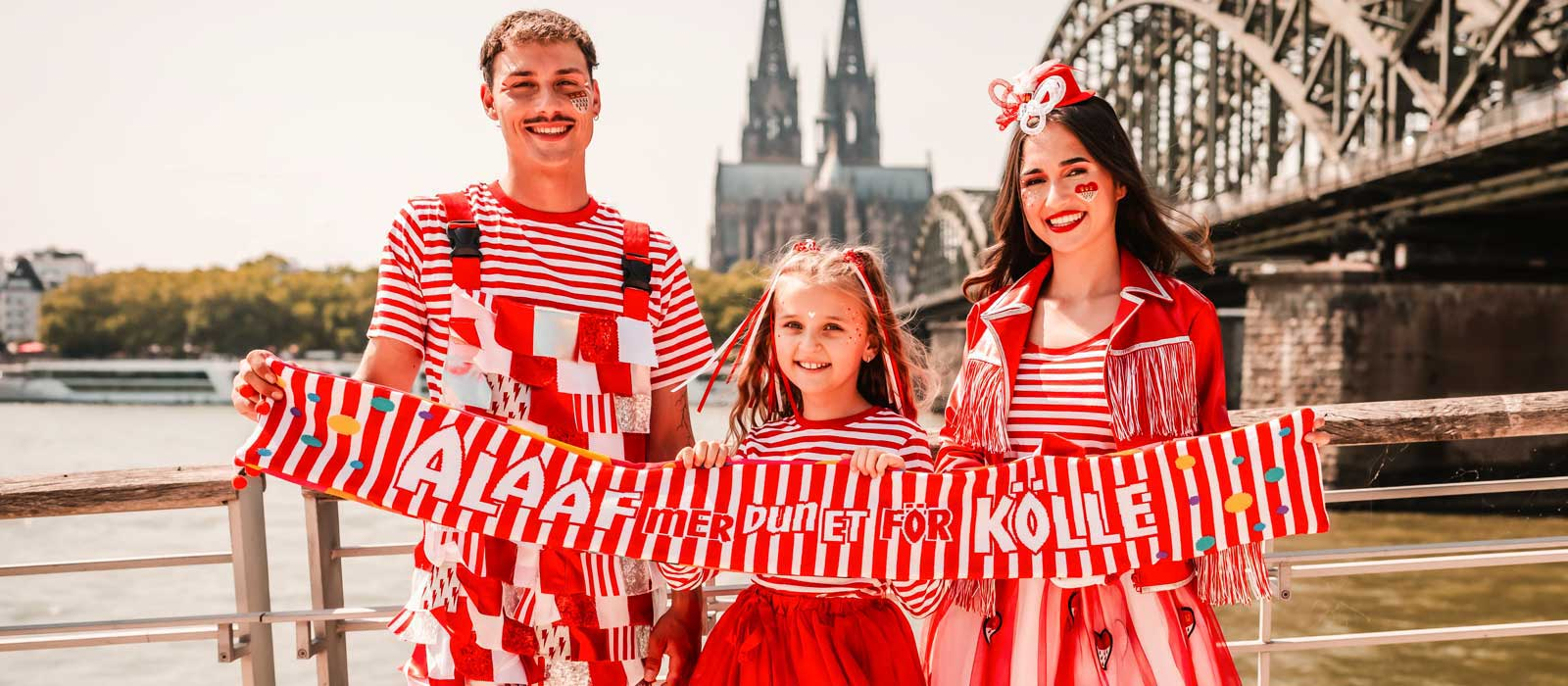 3 Personen vor der Kölner Skyline mit dem Kölner Mottoschal 2025/2026 und in rot-weißen Kostümen
