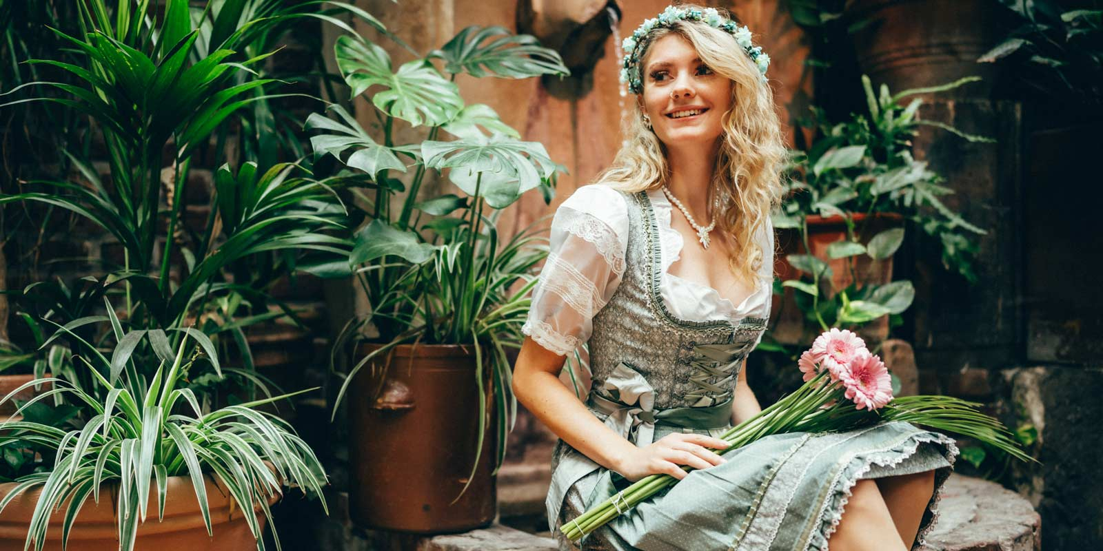 Eine Dame in mintfarbenem Dirndl sitzt am Brunnen mit Blumen in der Hand