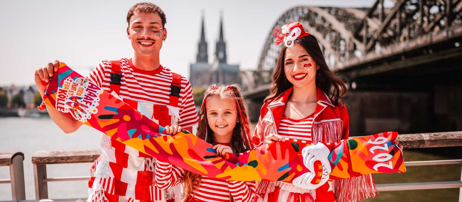3 Personen vor der Kölner Skyline mit dem Kölner Mottoschal 2025/2026 und in rot-weißen Kostümen